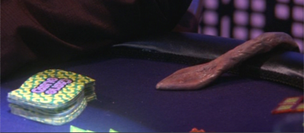 close-up of a table with a pack of cards on it, and a pink tentacle with a spade-shaped tip reaching toward them