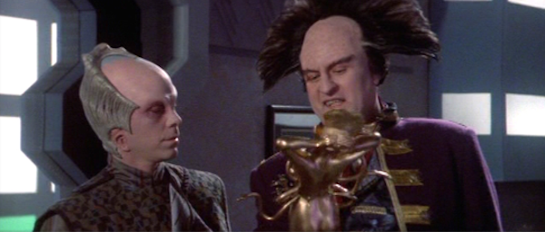 Londo Mollari, a humanoid man in a Napoleonic uniform, with an enormous axe-shaped hairdo resembling a mohawk turned 90 degrees, is showing Lennier a small golden statue. Lennier is a humanoid man with a crest of bone around the back of his bald head. The statue is a naked humanoid figure with 6 tentacles sticking out of its torso.