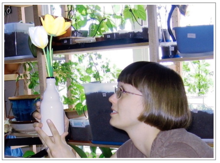 picture of me in my old apartment in Minneapolis, holding up a vase of white and yellow tulips in front of my large shelving unit full of potted plants