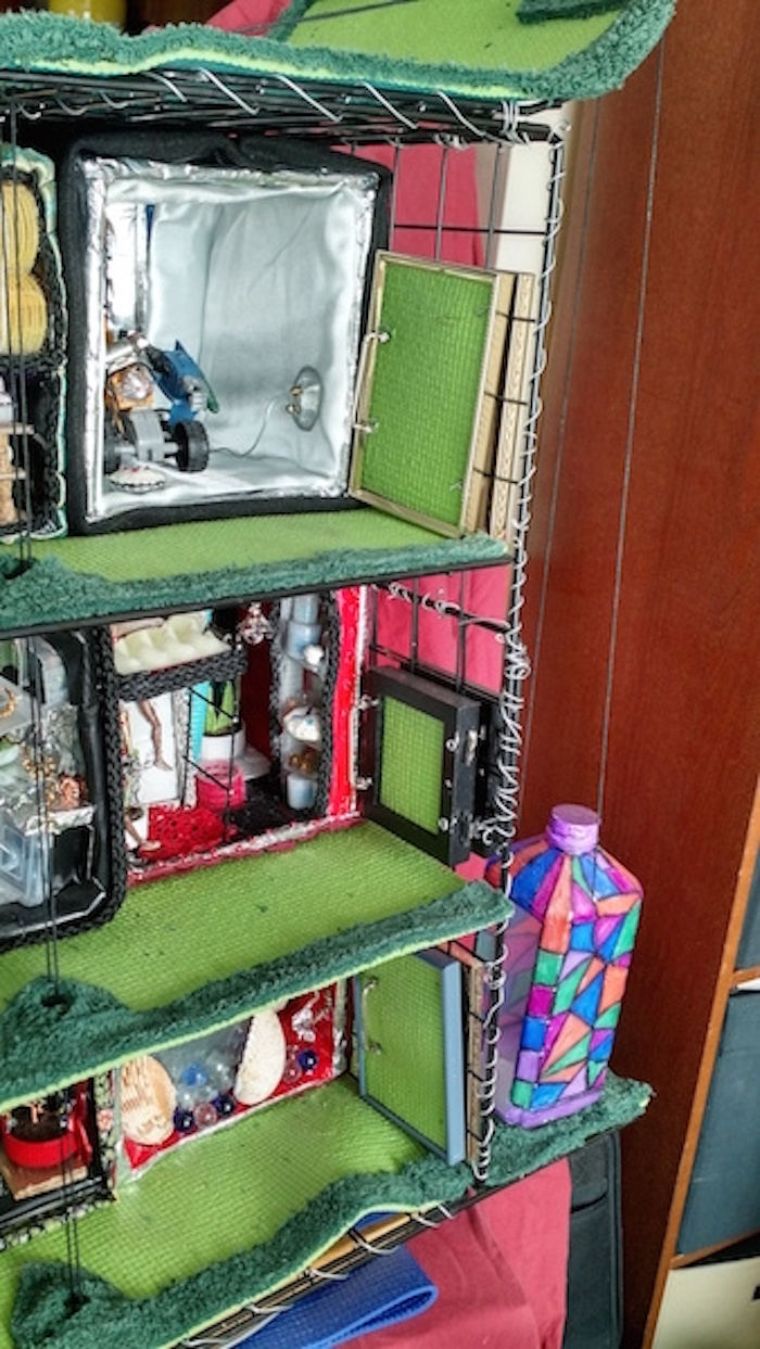 Another still photo of half-scale model of 6 apartment cubes on 3 stories of wire shelving. Same as previous one but centered more on the side with the elevator, made of a colorfully-decorated plastic bottle on strings.