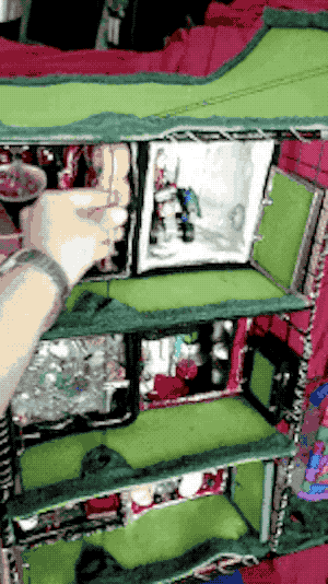 Animated GIF of a half-scale model I built of 6 apartment cubes on 3 stories of wire shelving, two to each floor. A colorfully-painted elevator made from a plastic bottle travels up and down, as my hand pulls cords connected to it.