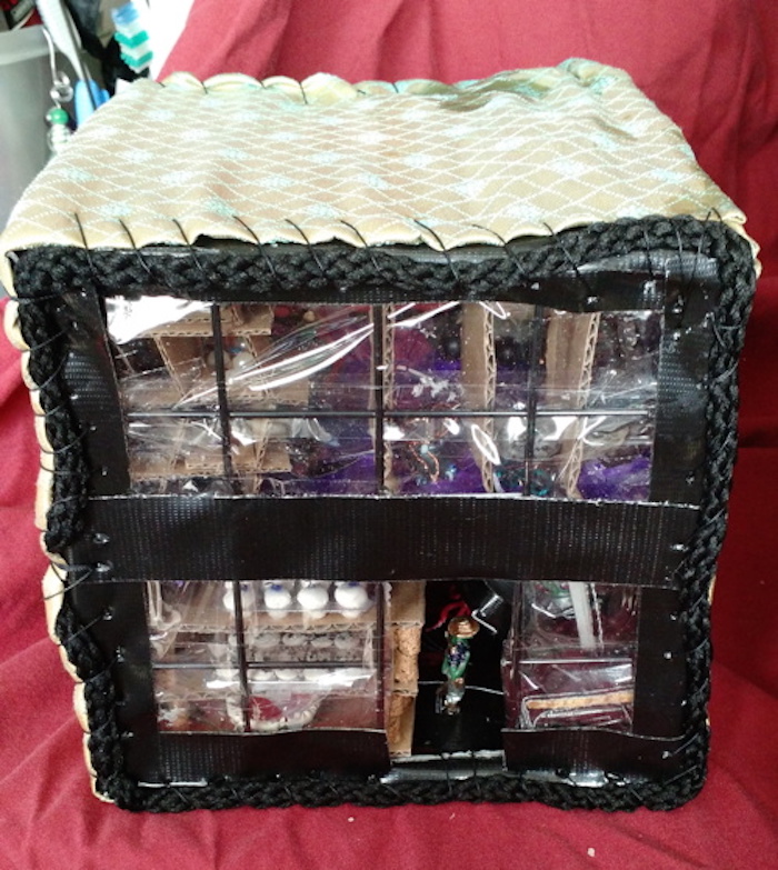 Photo of the Baking Cooperative mockup cube, removed from the shelving, seen from the closed outer side. Wall is made of transparent windows. Shelf of pastries can be seen through windows on first floor, beside an open door where a small green humanoid is visible. The roof and sides of the cube are wrapped in a golden, patterned fabric, sewn to a black rope that frames the perimeter of the windowed side.