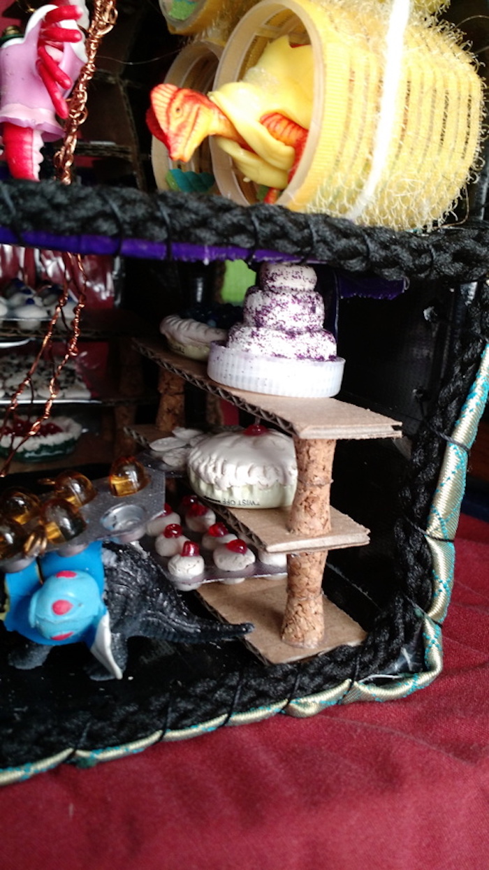 A view of the left side of the Baking Cooperative mockup cube, showing the hind end of the dinosaur carrying the egg carton, as well as the nearest shelf of pies, bead-topped muffins, cookies, and layer cake. Above them, part of the upper floor can be seen, with the ladder-climbing lobster, and the dinosaur going to bed in the yellow sleeping tube.