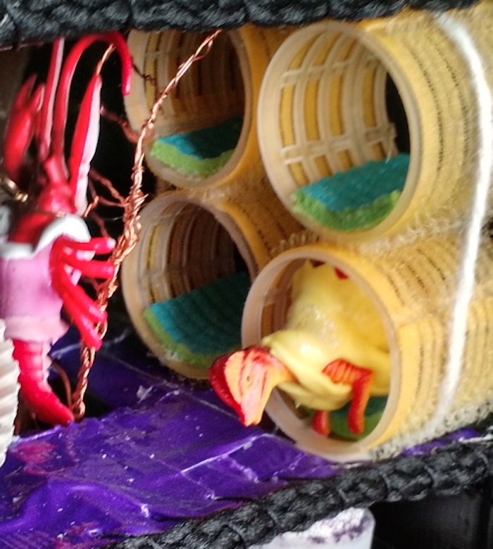 Close-up of the upstairs bedroom in the Baking Cooperative mockup cube, showing that the sleeping tubes in this version are made of yellow hair rollers lined with tiny pads of yoga-mat foam. A dinosaur, wearing a yellow rubber dress from a doll, sleeps in one of them, while a lobster in a similar pink dress climbs a ladder from the floor below.