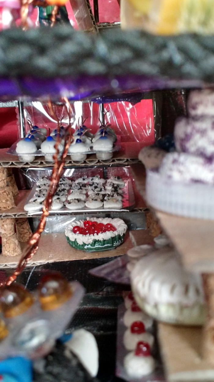 View of the display shelf of pastries against the window of the Baking Cooperative mockup cube. Top shelf is a muffin tin, made of a pill blister-pack, filled with clay muffins topped with blue beads. Second shelf is a tray of flat cookies covered in desiccant sprinkles; bottom shelf is an open-topped clay pie in a metal bottle cap, filled with red beads. More pies, cookies, and muffins are visible on another shelf in foreground, as well as a layer cake in a plastic bottle cap.
