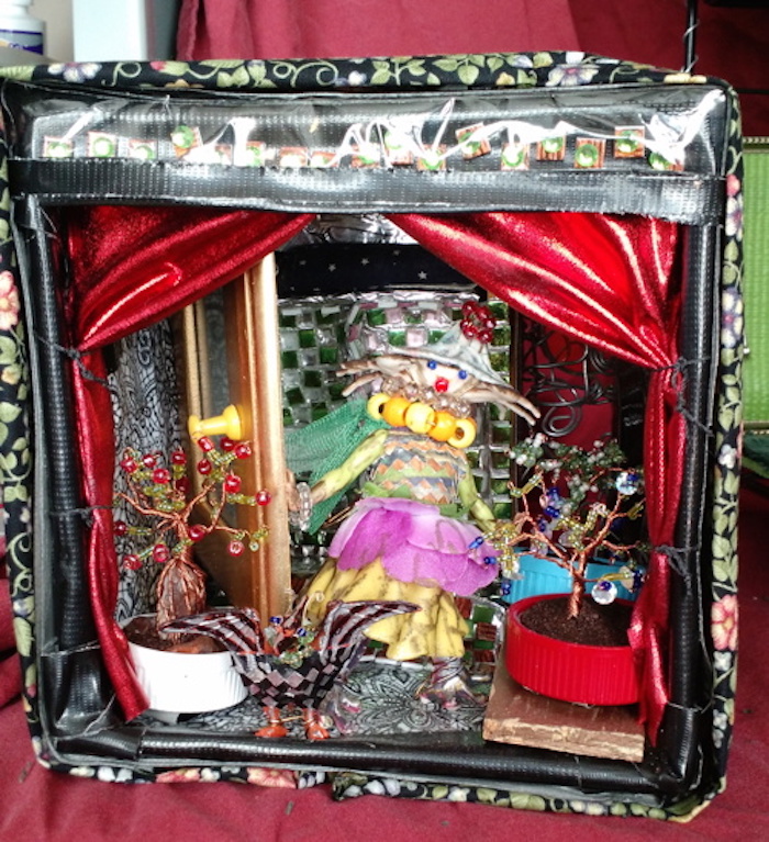 Photo of the sample mockup of the apartment, removed from the shelving, seen from the open side. Tree Mother is made from a small doll, dressed in pink and yellow doll skirts, whose head has been replaced with a tiny toy crab. Singer is a toy pterodactyl, who has been covered in feathers made from black and silver tape. Two small trees are visible in the foreground, made from wire and beads, and potted in plastic bottle caps filled with a mixture of glue and ground coffee. The bed-bath cubicle, made from a small wooden jewelry box and tiled on the inside with tiny white and green square tiles, is partly visible in the background.