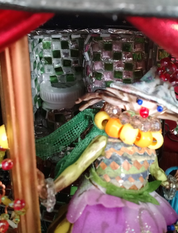 Photo of the sample mockup of the apartment, removed from the shelving, seen from the open side. This view focuses on Tree Mother's face, which has been adorned with beads to form eyes and a mouth. She wears a necklace of large orange and yellow beads, a green mesh scarf, and a hat made from a seashell decorated with a red bead flower. Behind her, the tiled bathroom cubicle can be seen more closely. The toilet is made from a short pill bottle, and is also covered in tiles to match the wall.