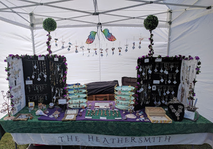 Photo of my table at the MN Renaissance Festival, outdoors under a canopy. Long table draped in green and white, with a tablecloth bearing the name THE HEATHERSMITH. 2 large rectangular stands with jewelry hanging all over them. Between them, handmade ornaments hang from a string between two dowels topped with leaf balls. 2 stands made of small turquoise plates hold handmade tiaras and crowns. Other smaller displays cover the table. A colorful pair of fairy wings hangs in the background. 