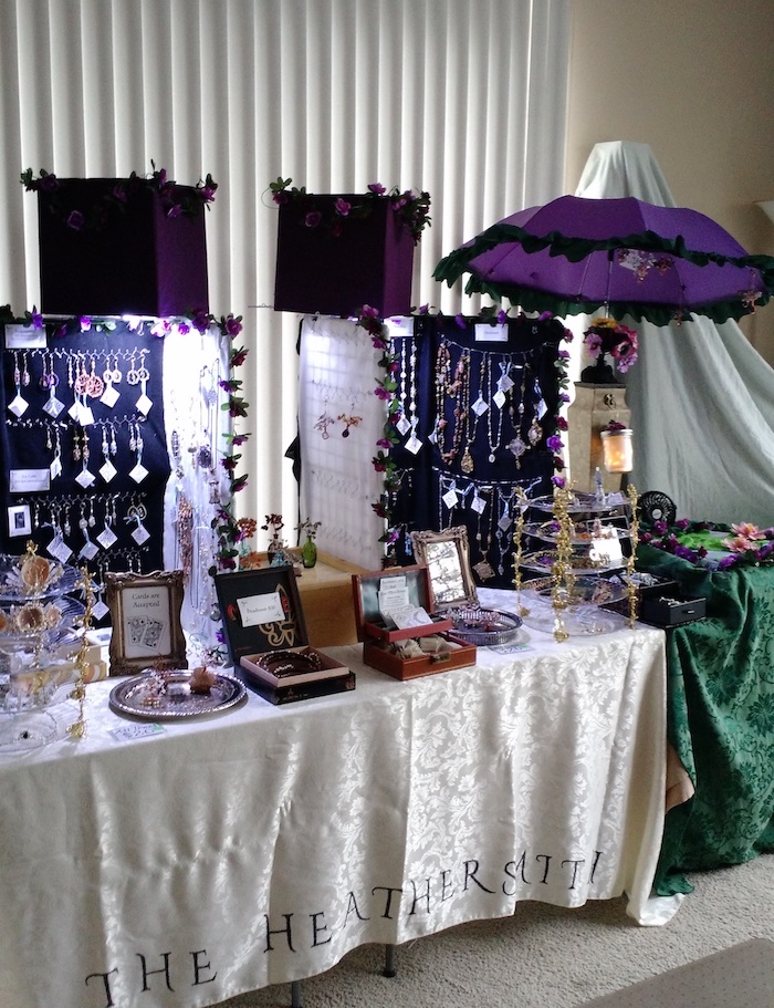Photo of a variation on the same setup, with battery-powered lights under square purple fabric cubes illuminating the displays. In this version, a purple umbrella with a green ruffle around the edge is set up with small ornaments hanging underneath it.