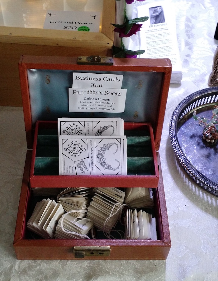 Photo of a small, open, 2-tiered jewelry box, with a sign on the inside of the lid saying Business Cards and Free Mini Books. The upper tier contains business cards for The Heathersmith. The lower tier is full of tiny paper books, each less than 2 inches square.